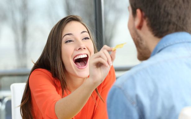 Woman in orange top laughing as she feeds potato crisps to a man in a blue shirt