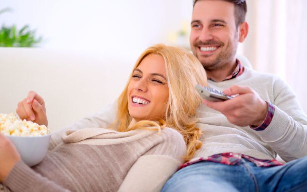 Brunette man and blonde woman lounging on safa and smiling at the tv while he holds a remote control